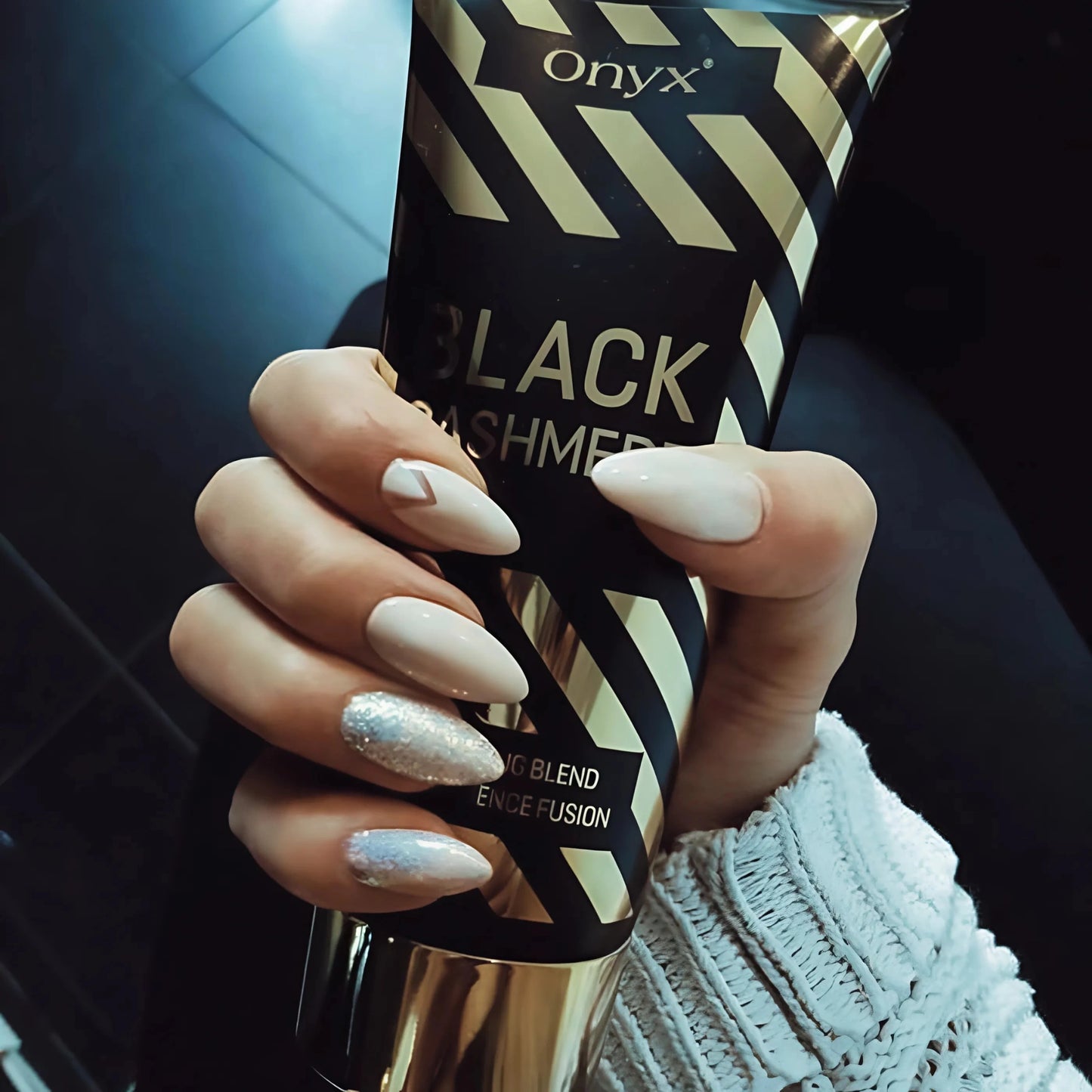 Hand mit hellen, glitzernden Nägeln hält Onyx Black Cashmere Bräunungscreme, eleganter Kontrast zu dunklem Hintergrund
