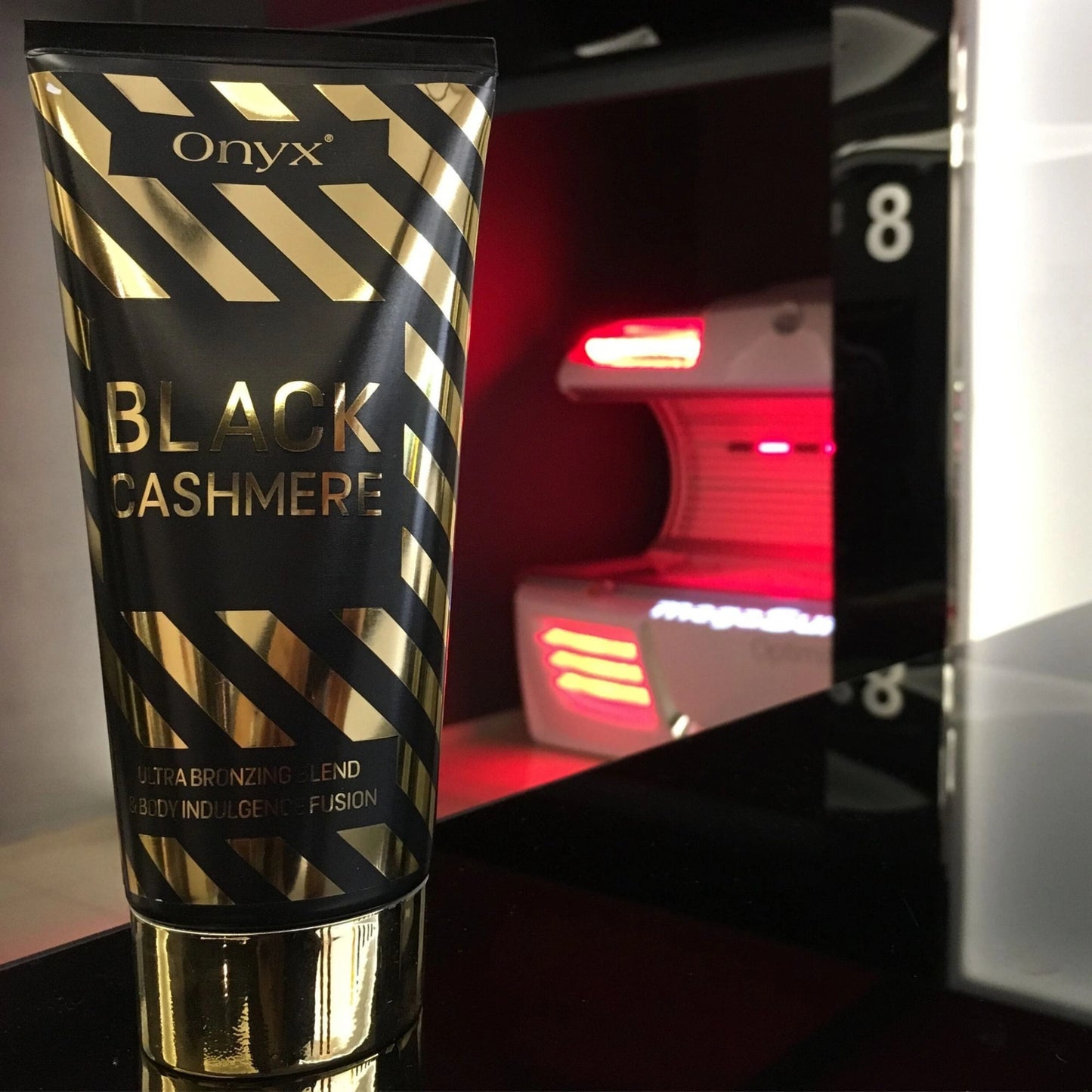 Onyx Black Cashmere Bräunungscreme steht auf glänzender Oberfläche vor einer modernen Sonnenliege mit rotem Licht