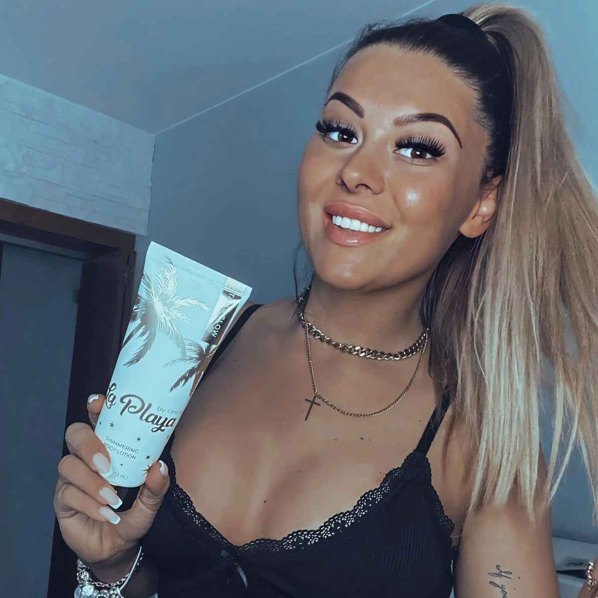 Frau mit gebräunter und glänzender Haut hält Onyx La Playa Tan & Glow Körpercreme in der Hand und lächelt