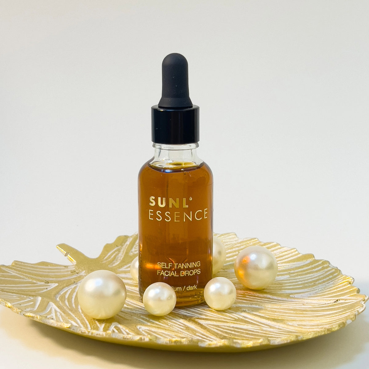 SUNL ESSENCE Selbstbräunungsdrops auf goldenem Blattteller mit dekorativen Perlen arrangiert