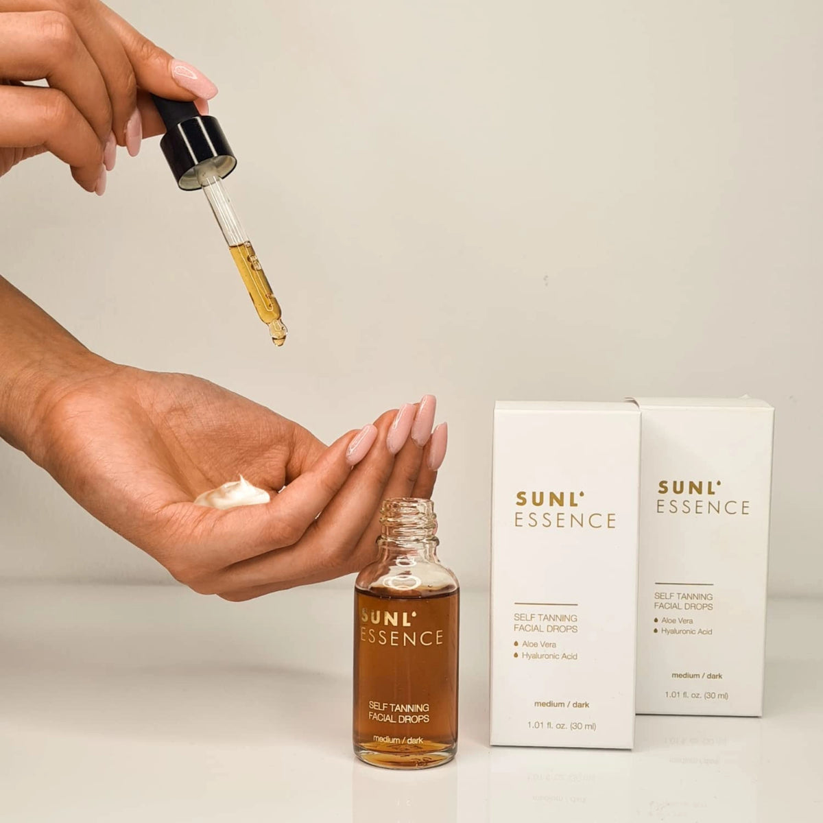 Hand mit rosafarbenen Nägeln träufelt SUNL ESSENCE Selbstbräunungsdrops auf Creme, Verpackung im Hintergrund sichtbar