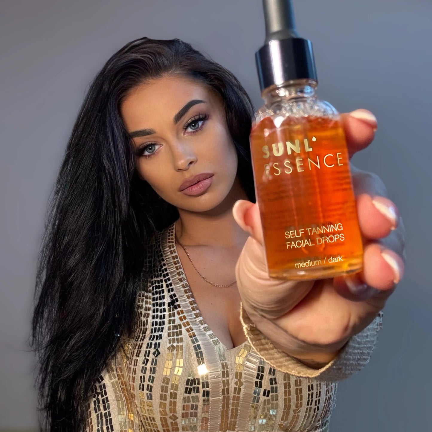 Frau mit langen dunklen Haaren und goldenem Paillettenkleid hält SUNL ESSENCE Selbstbräunungsdrops in die Kamera
