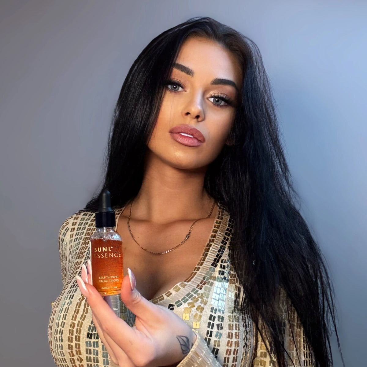Frau im goldenen Kleid zeigt geöffnete Flasche SUNL ESSENCE Selbstbräunungsdrops mit neutralem Hintergrund