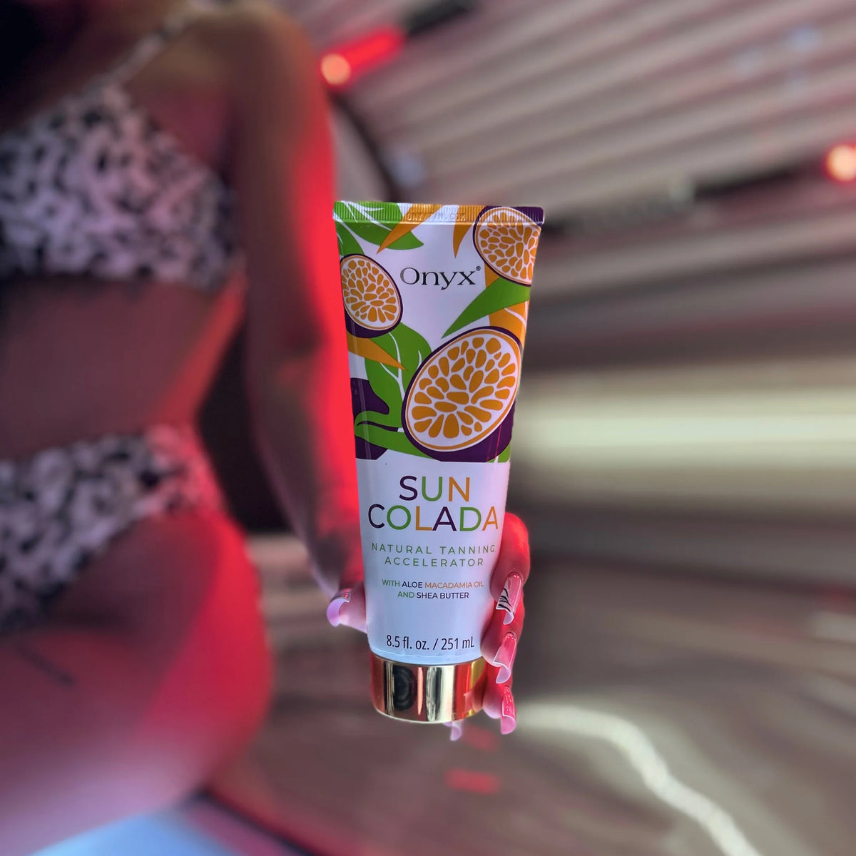Eine Hand hält die Tube Onyx Suncolada Natural Tanning Accelerator vor einer geöffneten Sonnenbank mit roter Beleuchtung.