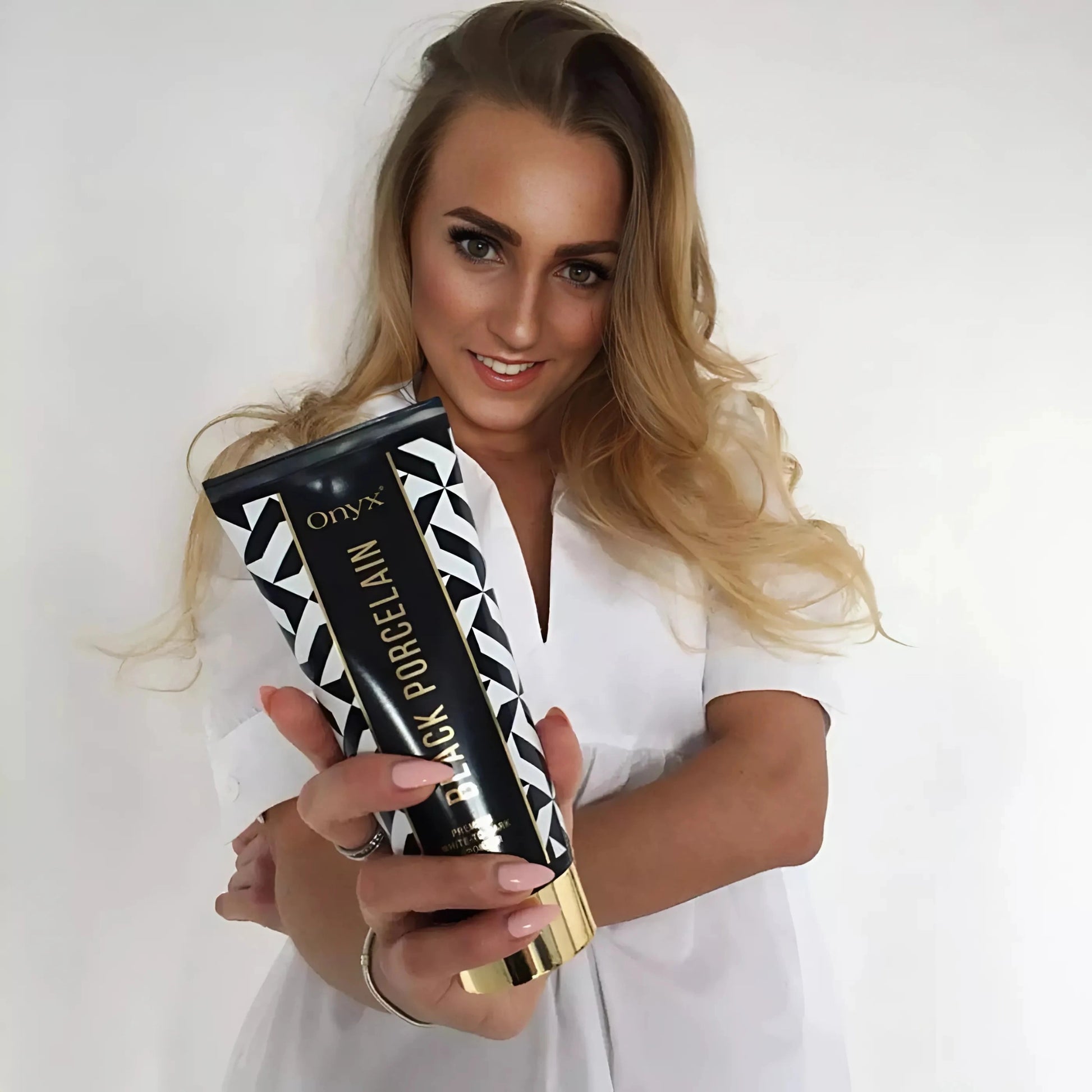 Blonde Frau in weißem Oberteil zeigt Onyx Black Porcelain Bräunungscreme vor heller Wand