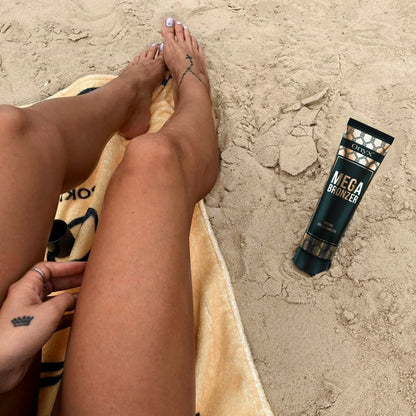 Onyx Mega Bronzer Tube im Sand neben braun gebrannten Beinen, zeigt natürliche Bräune nach Anwendung am Strand