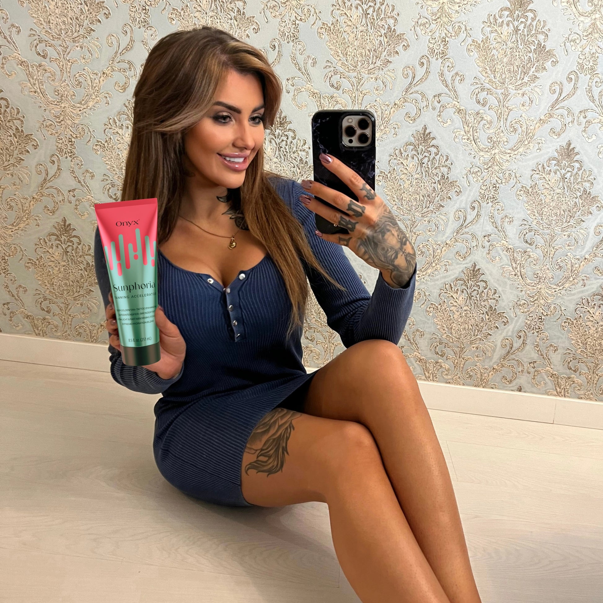 Frau mit Tattoos hält Onyx Sunphoria Bräunungslotion beim Spiegelselfie