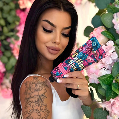 Eine Frau mit tätowiertem Arm hält die farbenfrohe Tube Onyx Tattoo Rose Bronzer vor einer dekorativen Blumenwand in Rosa- und Grüntönen.